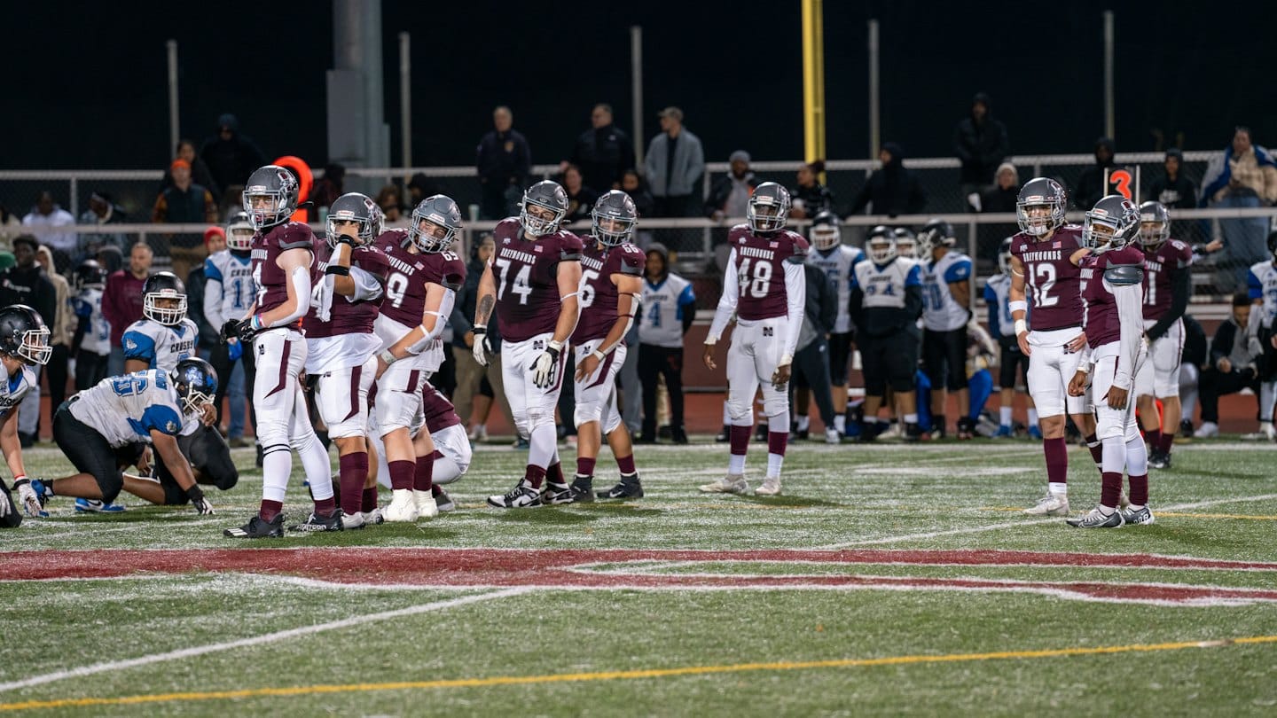 Football players in team uniforms stand on a turf field.