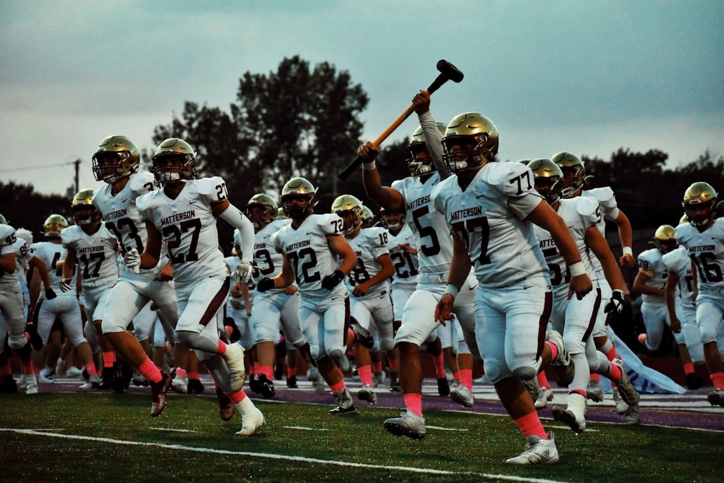 Young football players in white and gold uniforms run onto a field at dusk.